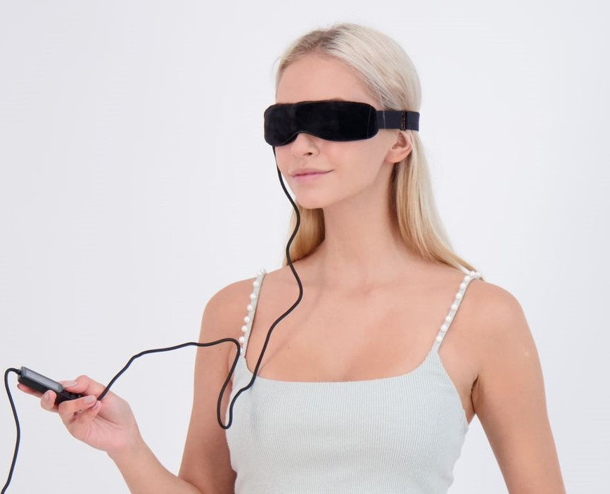 Blonde woman wearing black heated dry eye mask and holding remote control device.