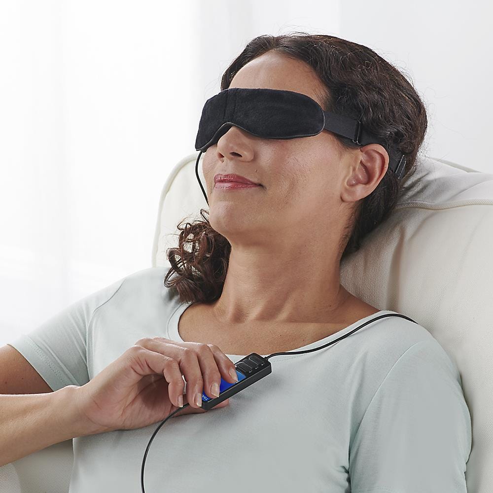 Middle-aged woman relaxing in a recliner wearing the Wizard Research heated eye mask while adjusting the settings with a wired remote control